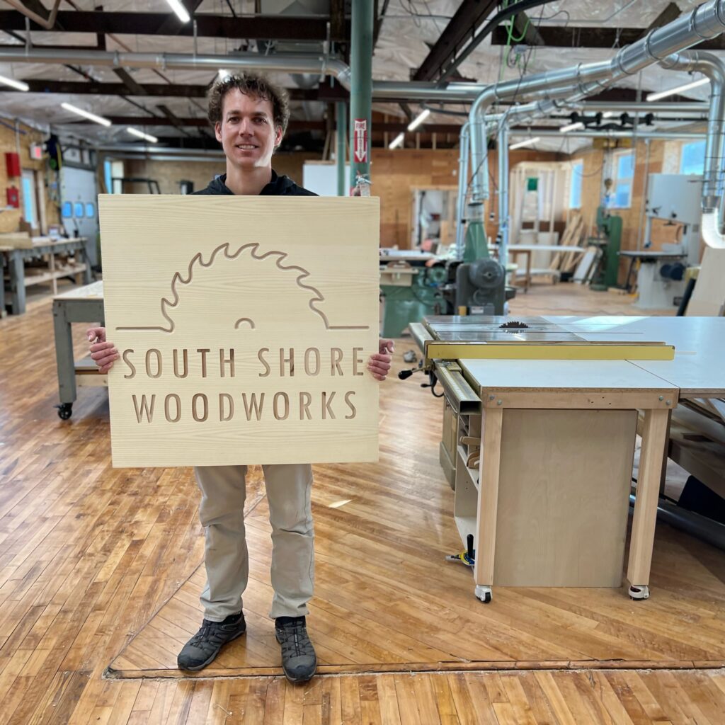 Sandy holding a sign that says South Shore Woodworkers