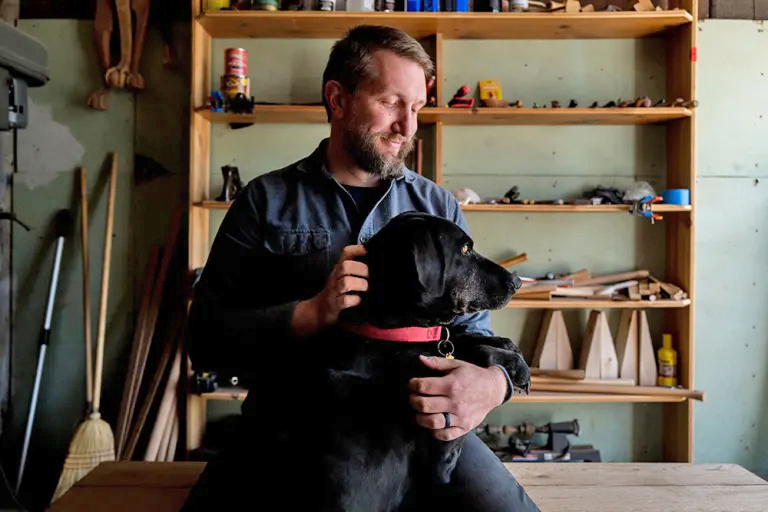 Andy holding his dog, a black lab, with shelves of tools behind him