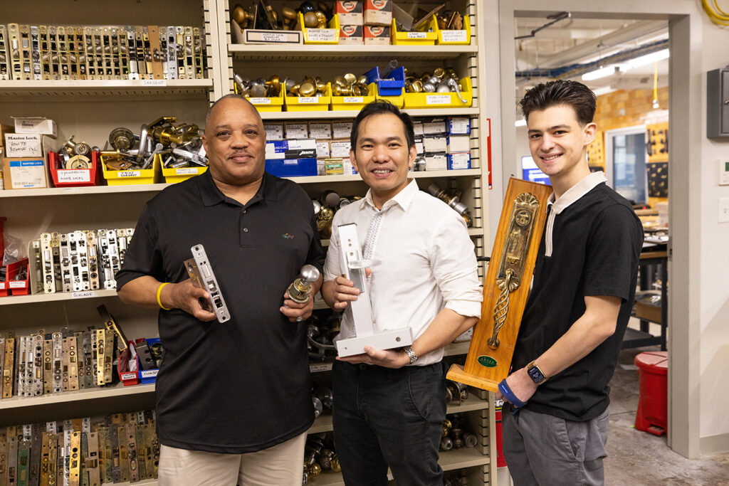 Three locksmithing students each holding hardware, in front of shelves